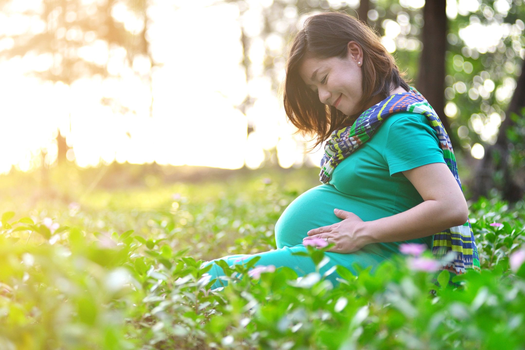 Pregnant woman sitting in a field of flowers and sunshine holds her belly and smiles at it.She's wearing a teal dress and a multi-colored scarf.