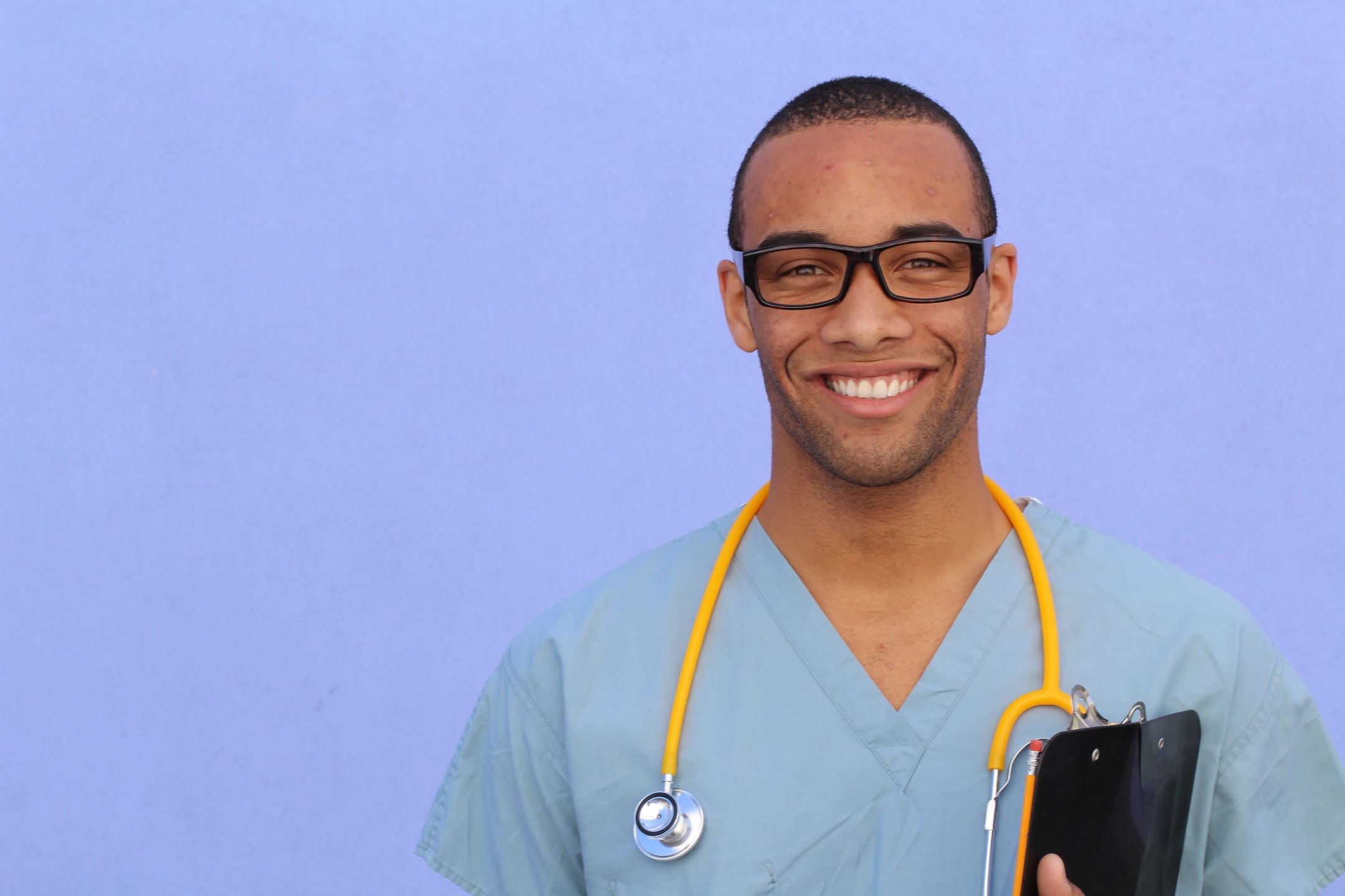 Healthcare worker wearing blue scrubs, glasses and a yellow stethoscope holding a clipboard and smiling at the camera