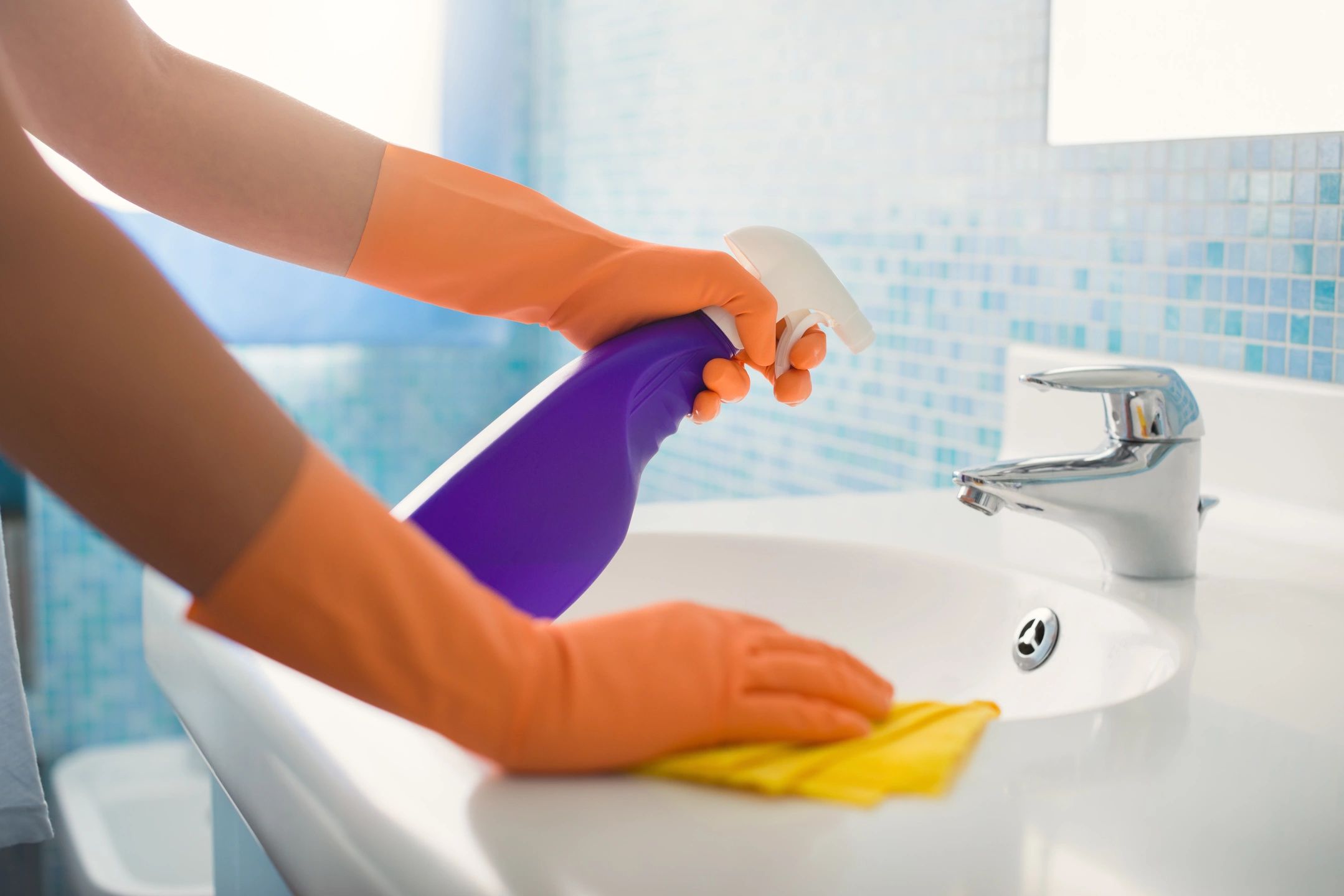 Person wearing rubber gloves cleans a sink with a spray bottle and a cloth