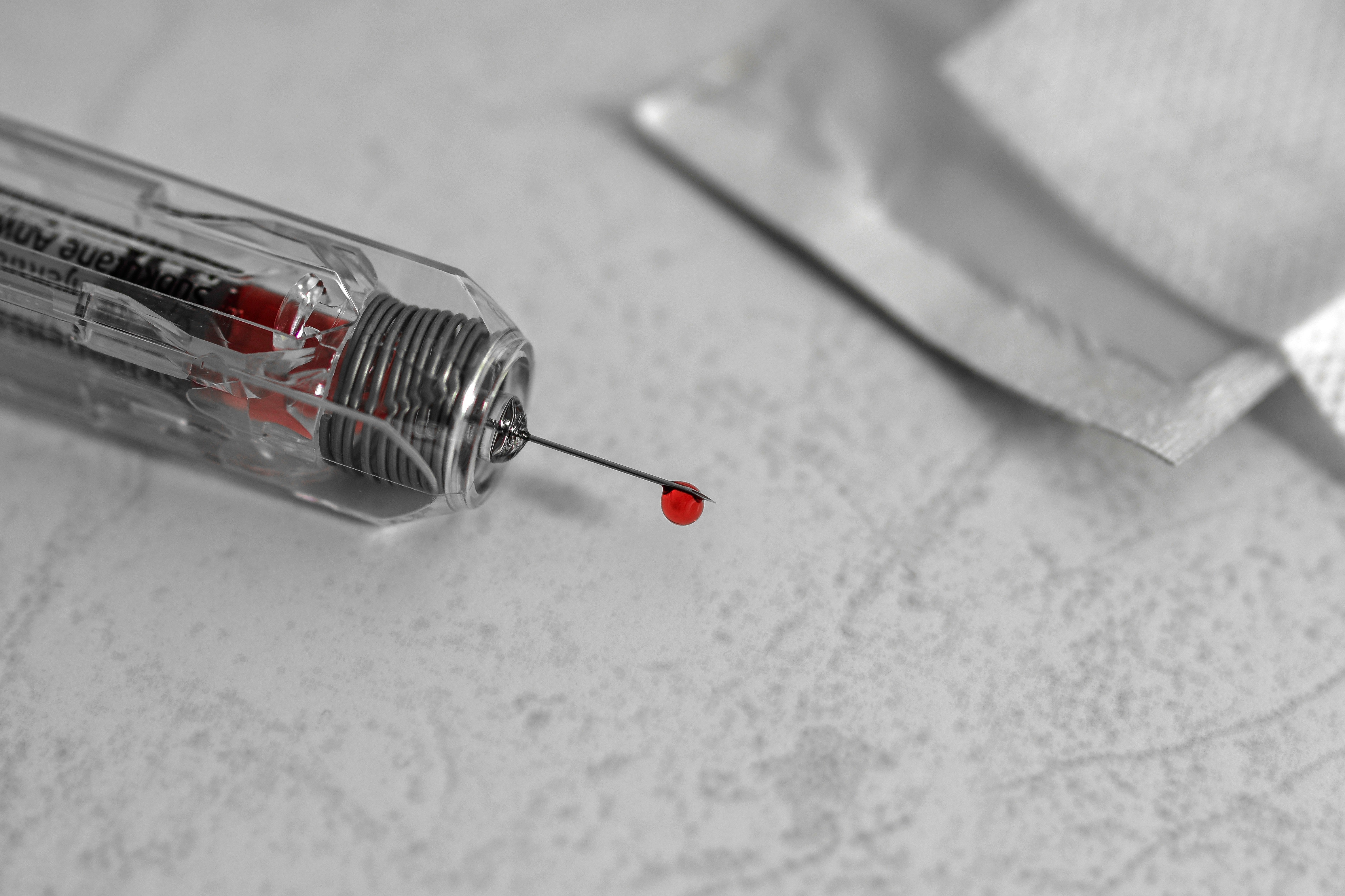 Close up of a blood drop on the end of a needle, which is attached to a syringe. The syringe is sitting on a hard grey surface.